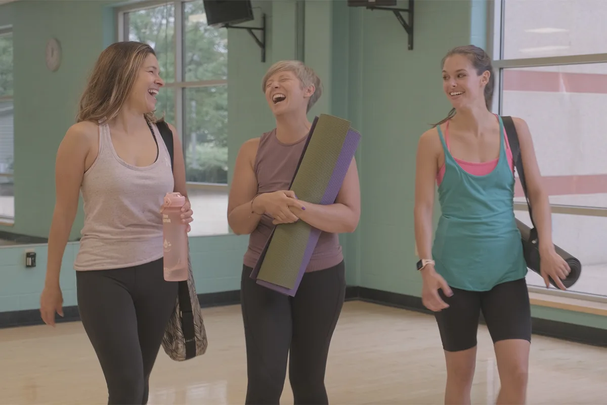 Three women in activewear laugh together while holding yoga mats and water bottles, suggesting a supportive gym atmosphere.