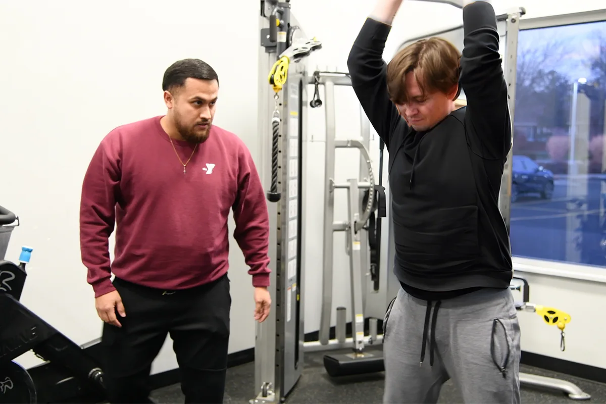 Trainer watches as young man lifts weights overhead in a gym setting.