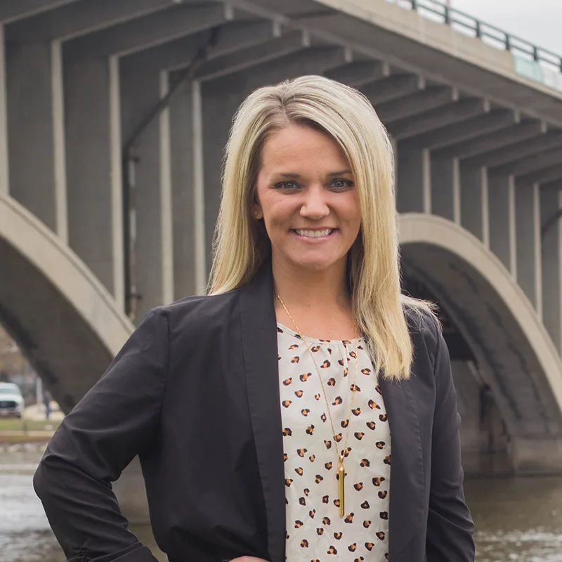 Smiling woman stands outdoors in front of a bridge, wearing a black blazer and patterned blouse.