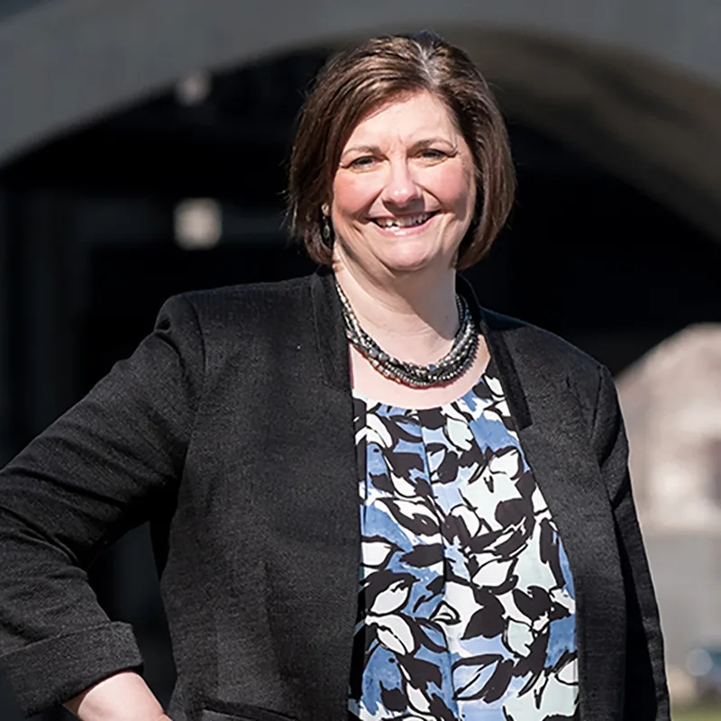 Smiling woman outdoors in professional attire with a black blazer and patterned blouse, posing confidently in front of a bridge.