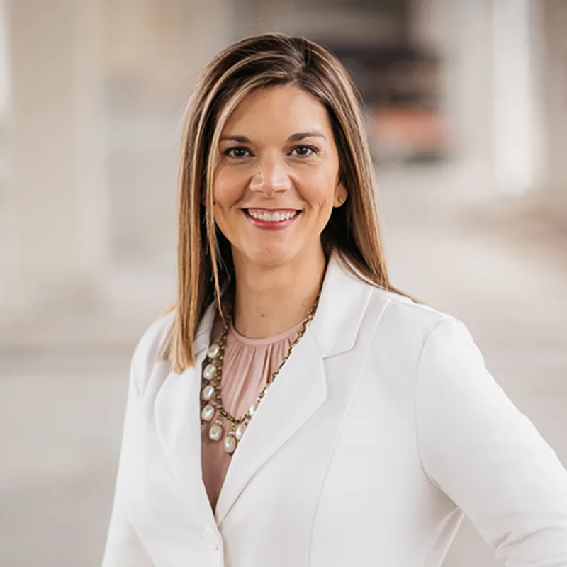 Smiling woman in a white blazer and statement necklace, standing confidently indoors with a blurred neutral background.