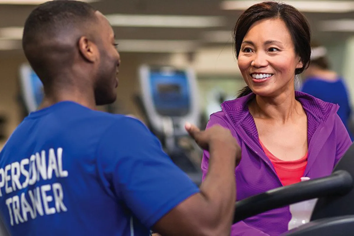 A personal trainer in a blue shirt speaks with a woman in a purple jacket, both smiling and engaged in conversation at a gym.