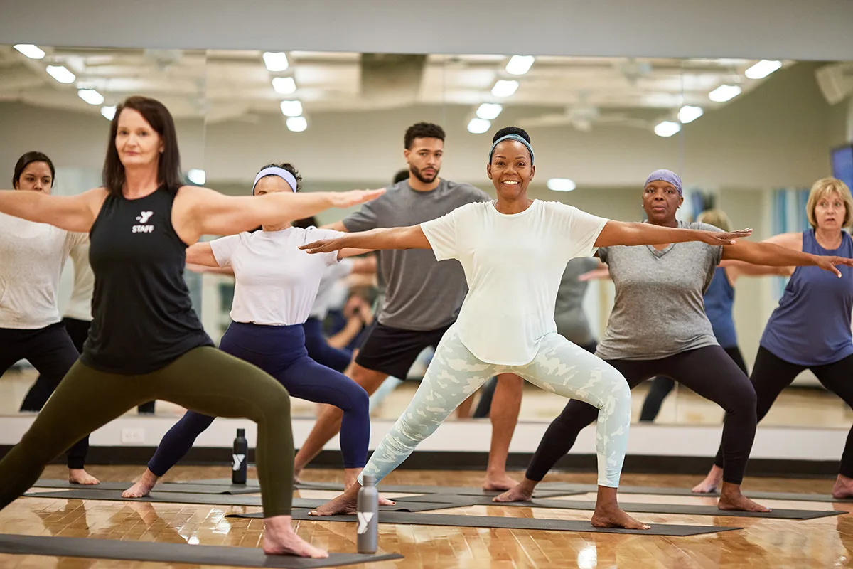 A diverse group of adults practicing yoga in a mirrored studio, holding Warrior II pose under the guidance of an instructor.