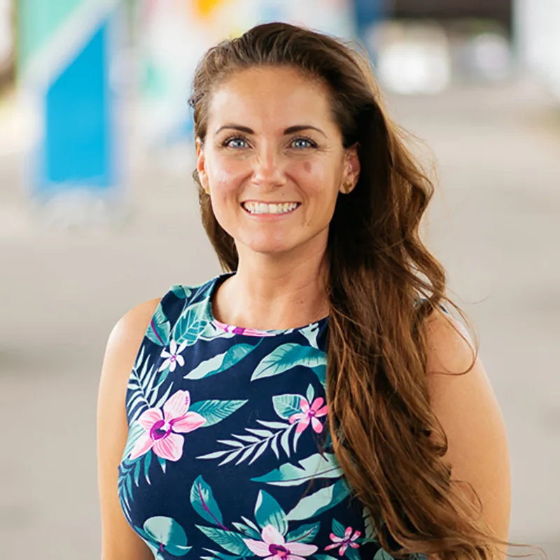 A woman with long brown hair smiles confidently while wearing a floral-patterned dress, standing outdoors with colorful murals in the background.