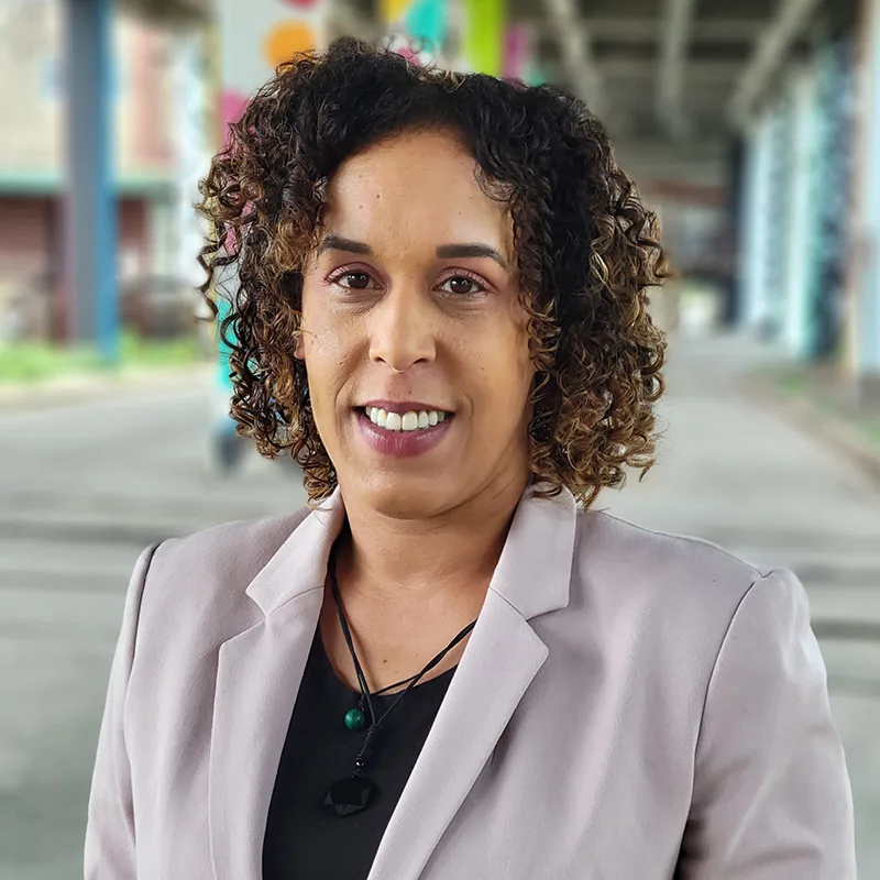 A woman with curly hair smiles while wearing a light blazer and dark top, standing outdoors with a colorful, urban backdrop.