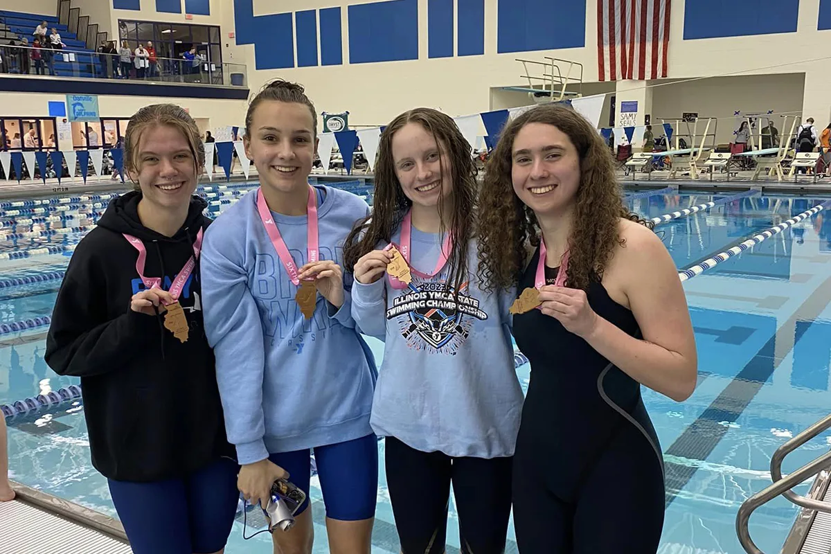 Four young female swimmers smile and hold medals by a pool after a competition.