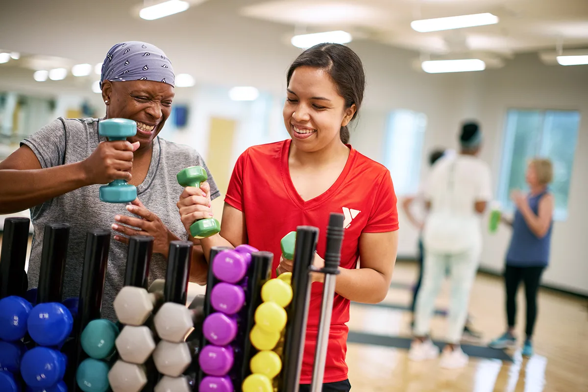 Two women smiling and selecting dumbbells in a gym, with a YMCA instructor helping in a bright exercise room.