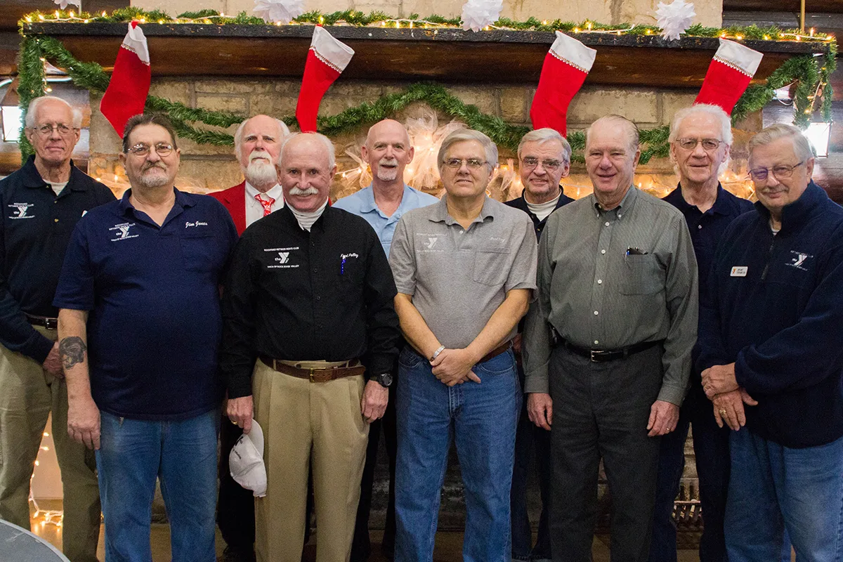 Group of men posing in front of a festive fireplace decorated with stockings, garlands, and holiday lights.