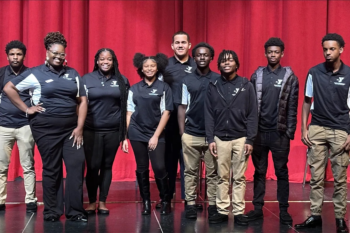 Group of young adults and their leader in matching YMCA shirts, standing on a stage in front of red curtains.