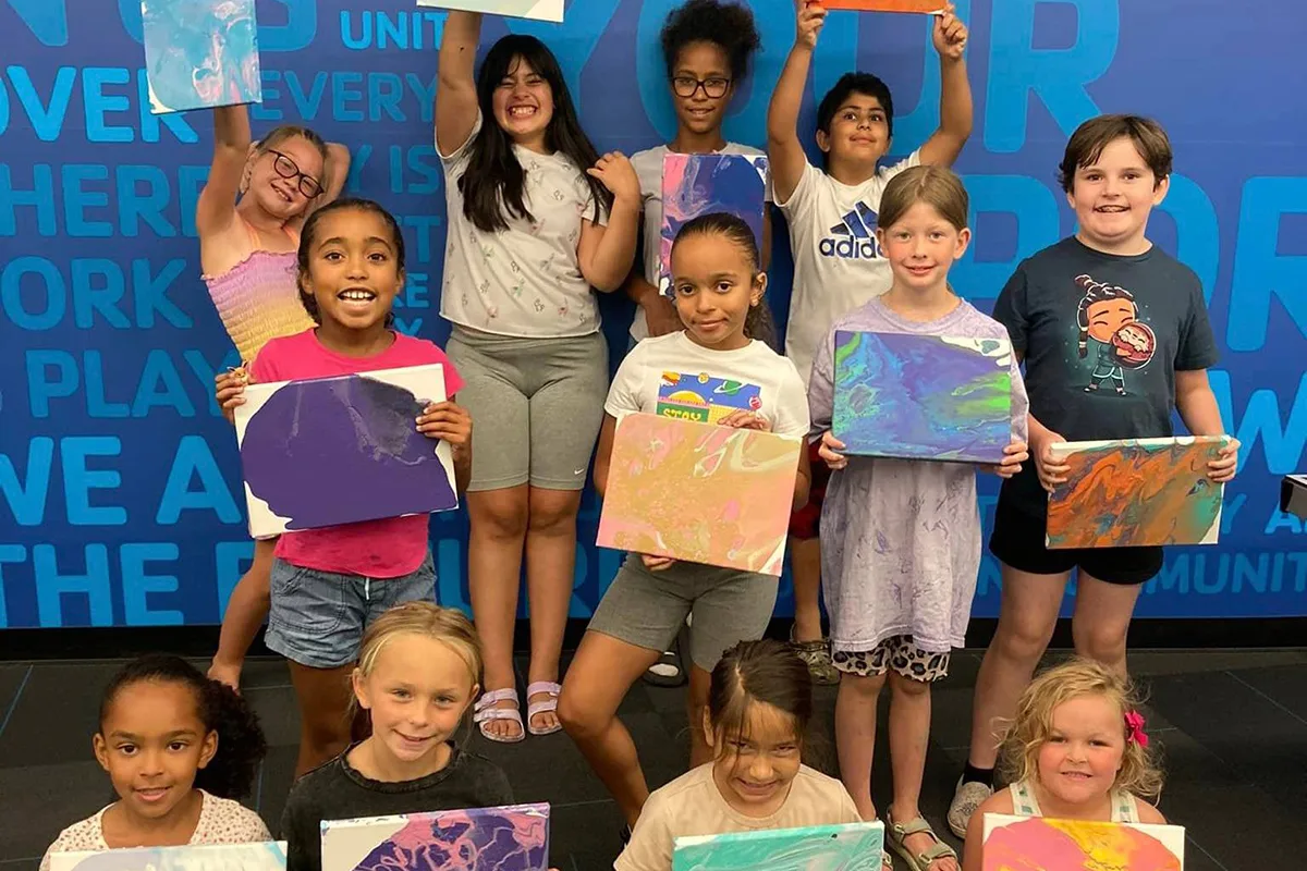 Group of children proudly displaying their colorful art pieces, smiling in front of a blue wall with inspirational words.