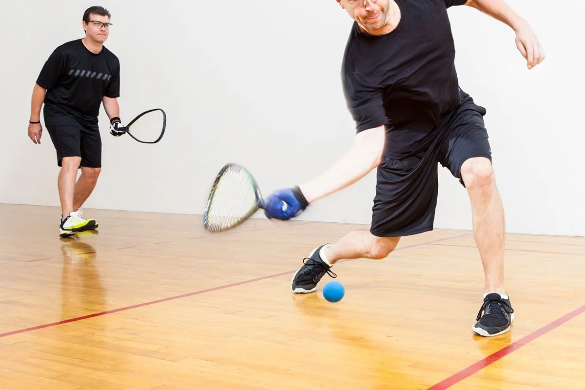 Two men playing racquetball in an indoor court, with one player lunging forward to hit the ball.