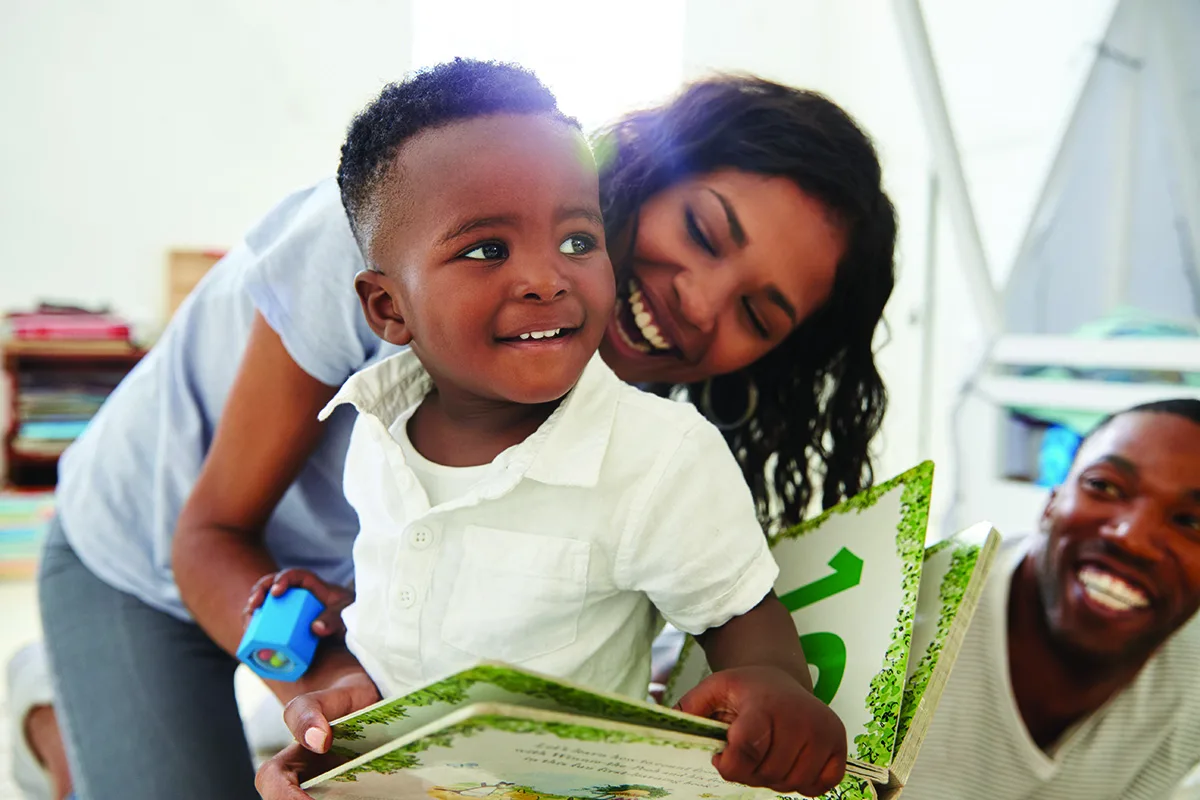 A young boy smiles while looking at a book, with two adults beside him smiling and engaging in the activity together.