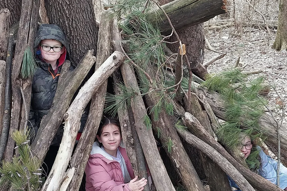 Three children smile from inside a makeshift shelter built with branches and pine boughs in a wooded area.