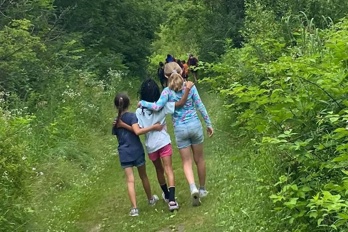 Three young girls walk along a forest trail with their arms around each other, surrounded by green foliage. A group of campers can be seen further up the path.