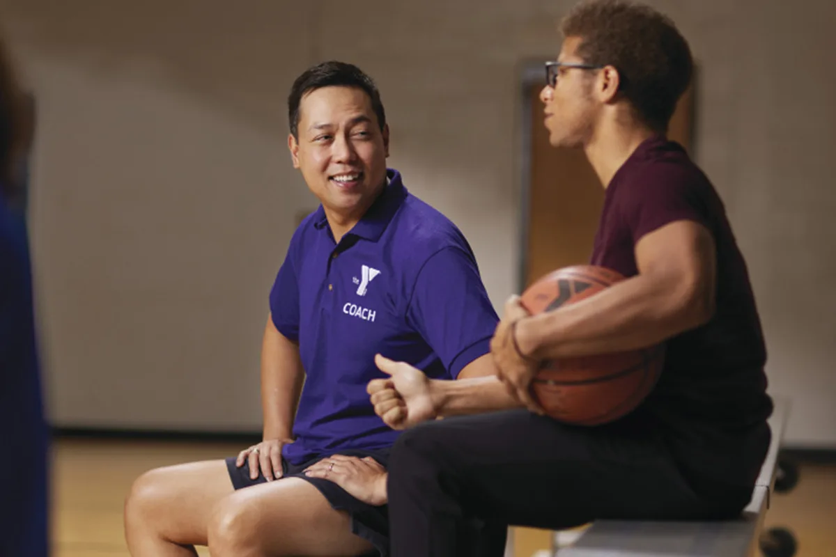 A basketball coach in a purple shirt smiles while talking with a young man holding a basketball on a gym bench.