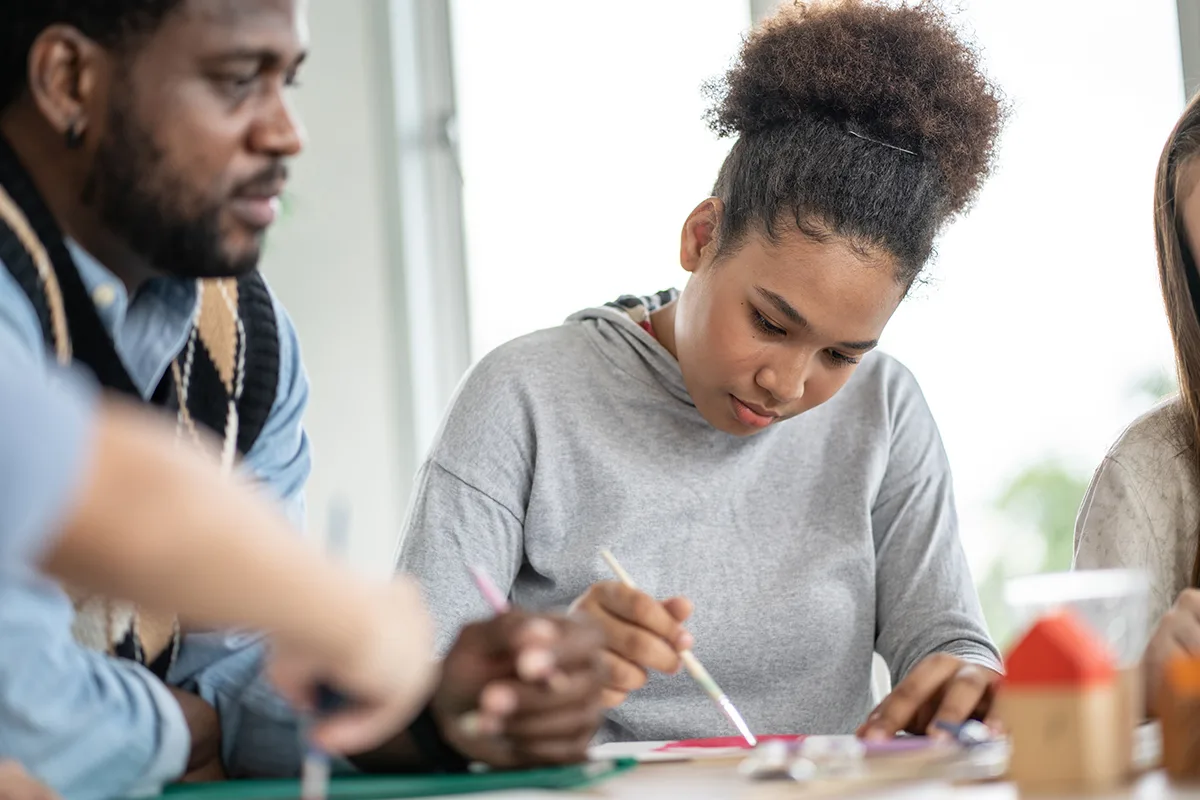 Young woman focused on a painting project in art class, with instructor observing closely.