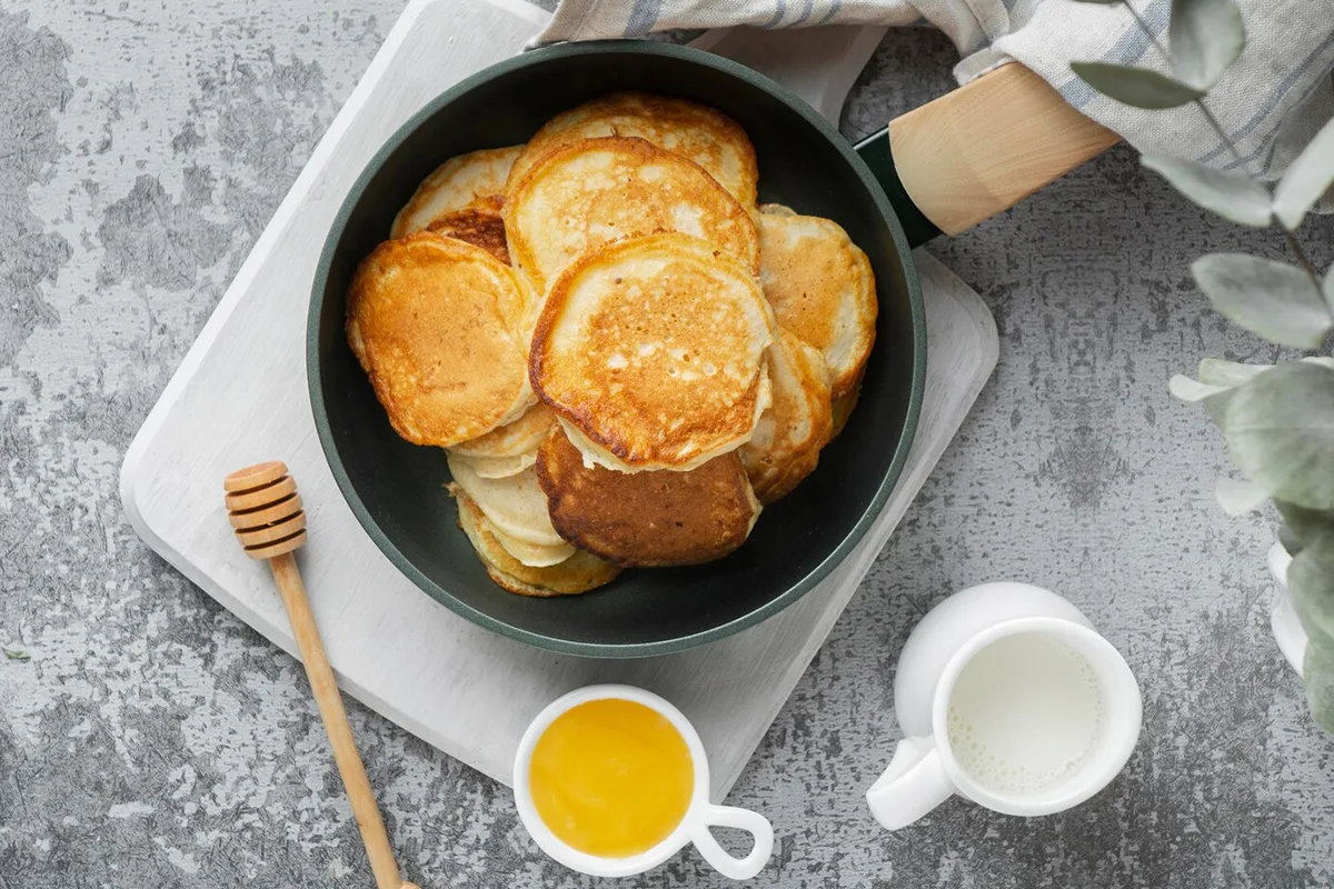 Top view of golden pancakes in a skillet with honey, butter, and milk on the side, set on a rustic gray background.