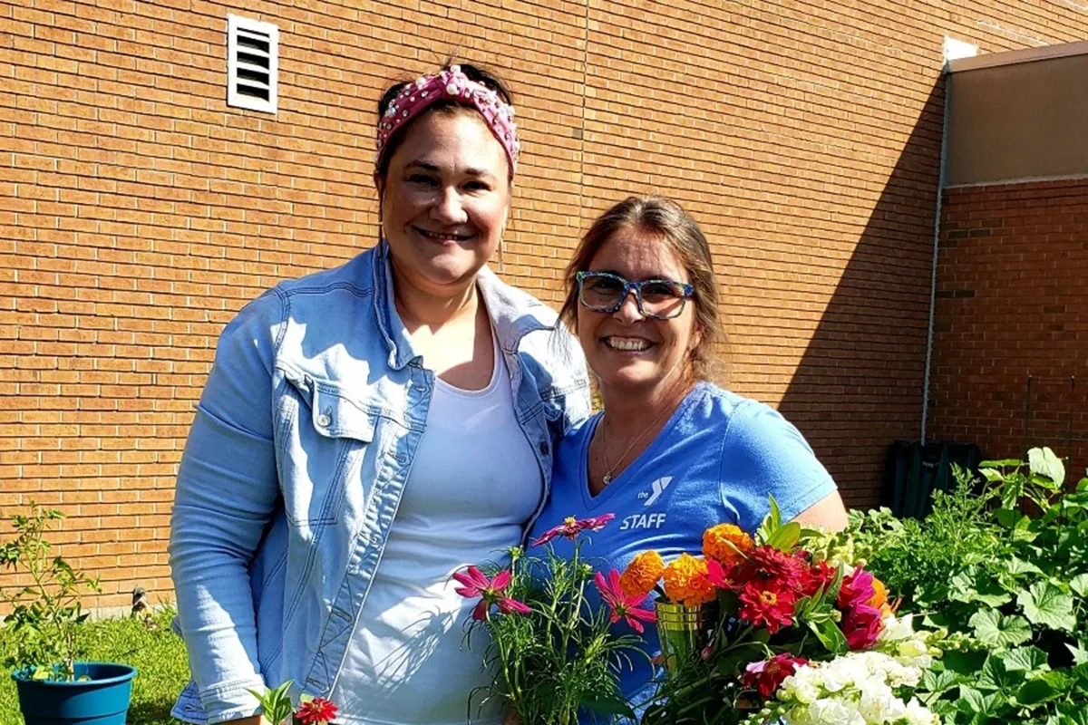 Two women smiling outdoors, holding freshly picked flowers near a garden with a brick building in the background.