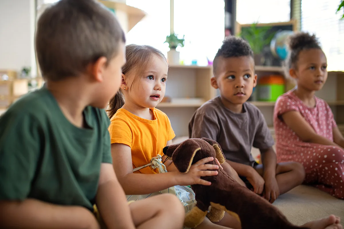 Children sitting together in a classroom, holding a stuffed animal.