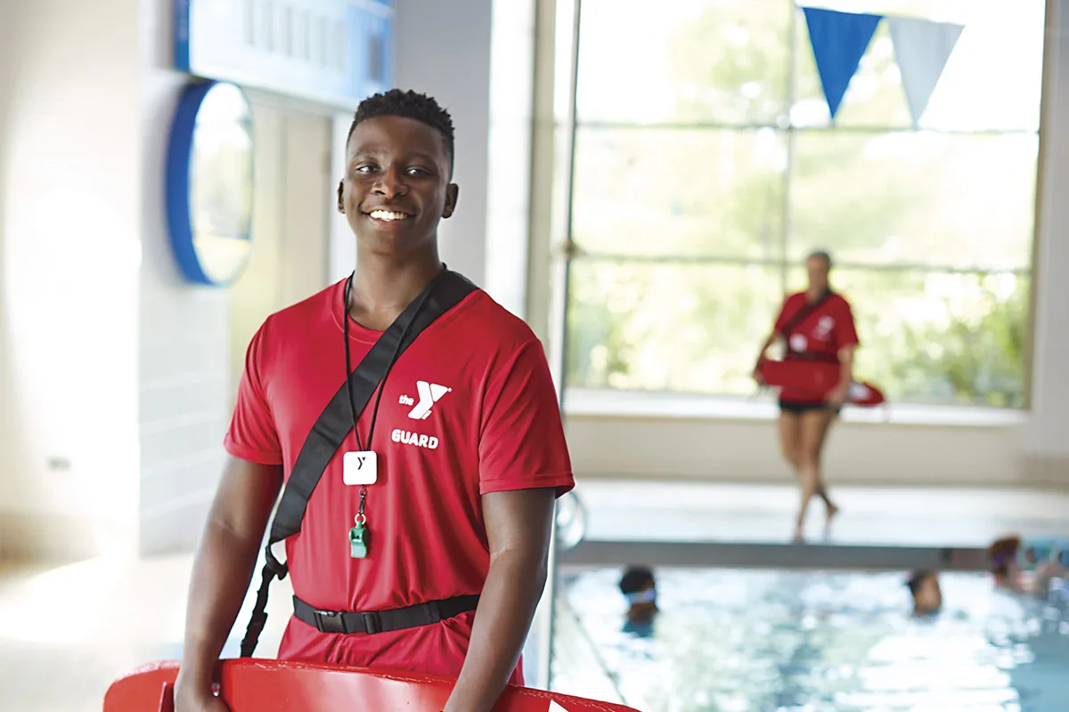 Young lifeguard in red uniform stands by an indoor pool, with swimmers and another lifeguard in the background.