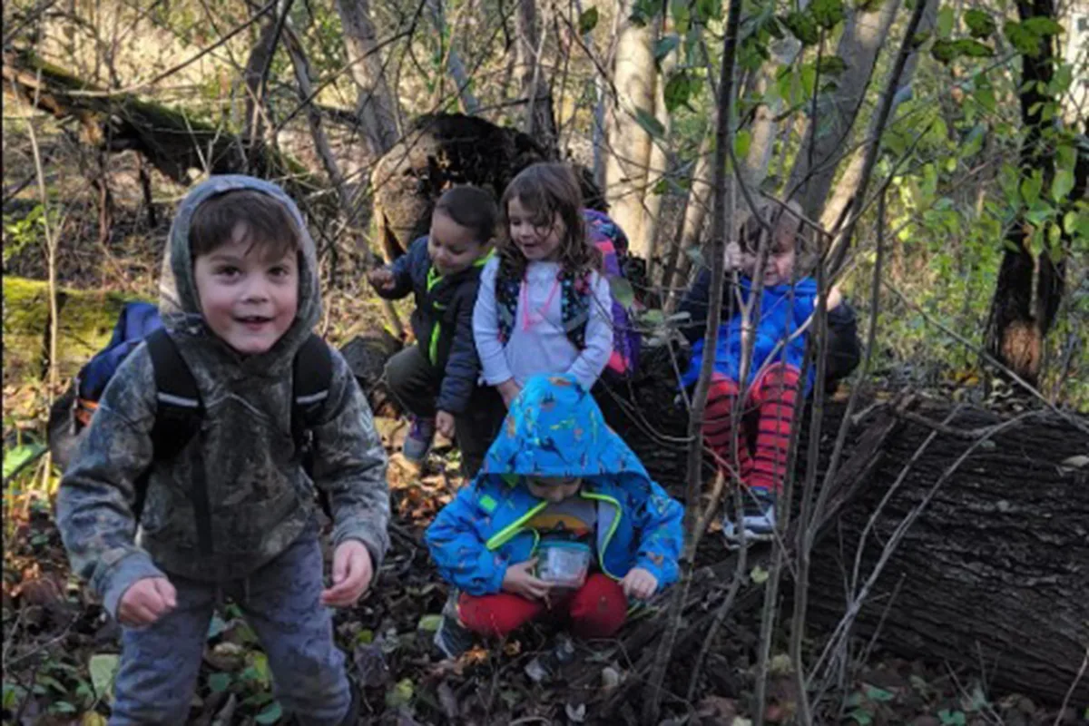 Group of young children wearing jackets and backpacks exploring a wooded area, surrounded by trees and branches.