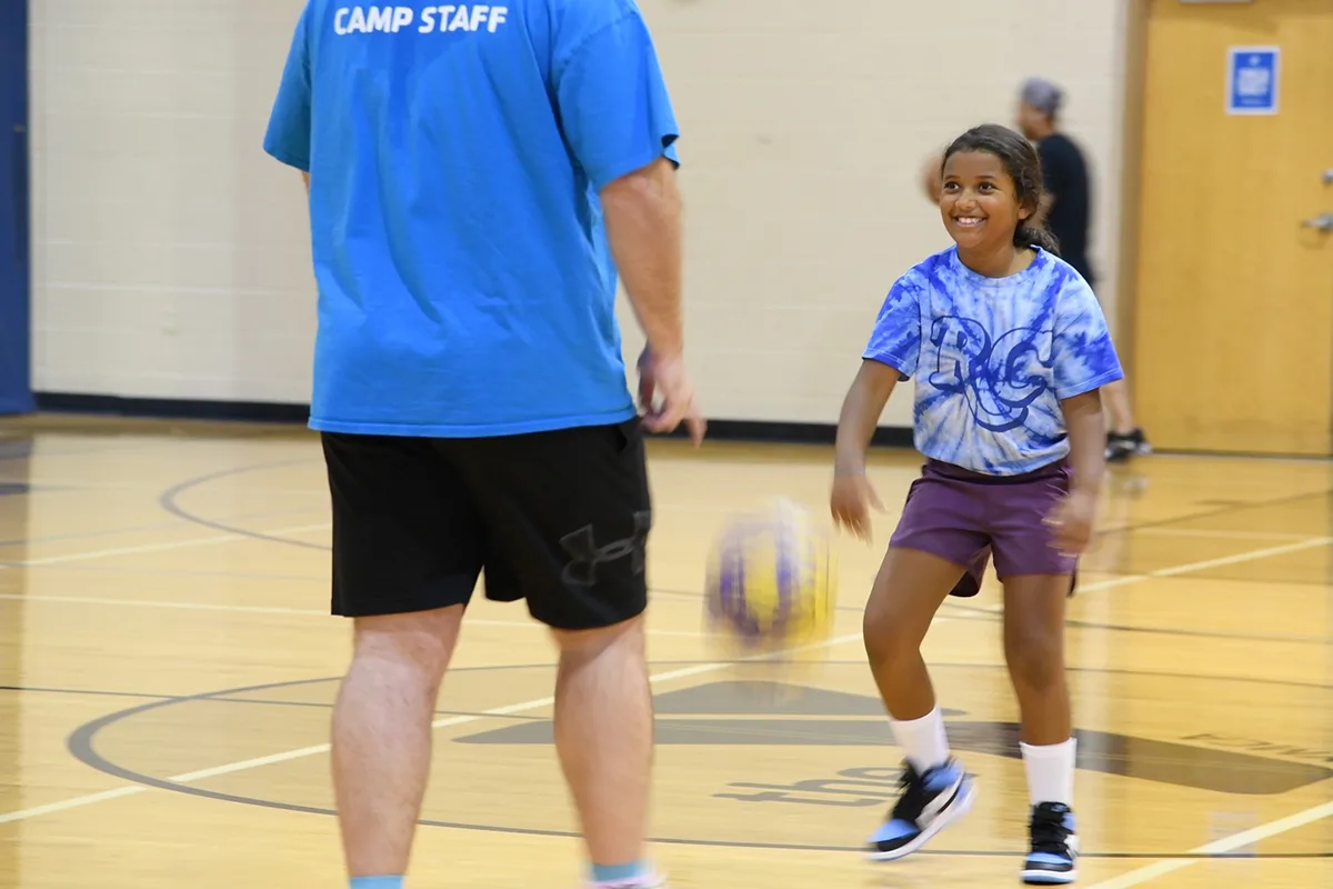 A young girl in a tie-dye shirt smiles while playing with a basketball in a gym.