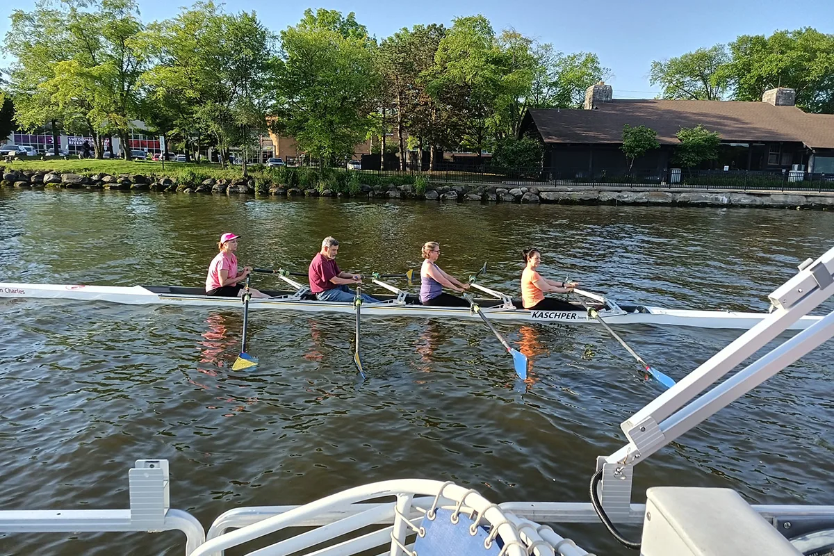 Four-person rowing team paddling on a calm lake near a tree-lined shore, with a nearby dock and building in the background under a clear blue sky.