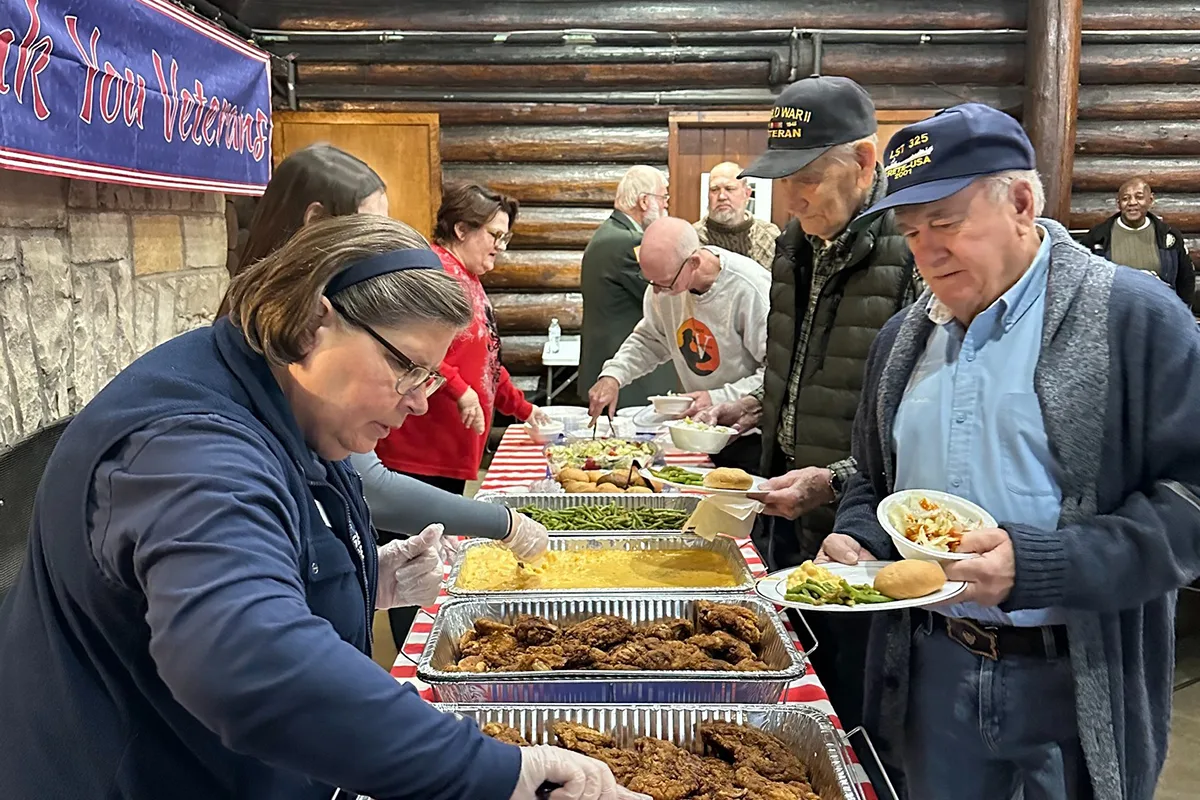 Veterans and volunteers at a buffet table, serving food under a 'Thank You Veterans' banner in a log cabin setting.