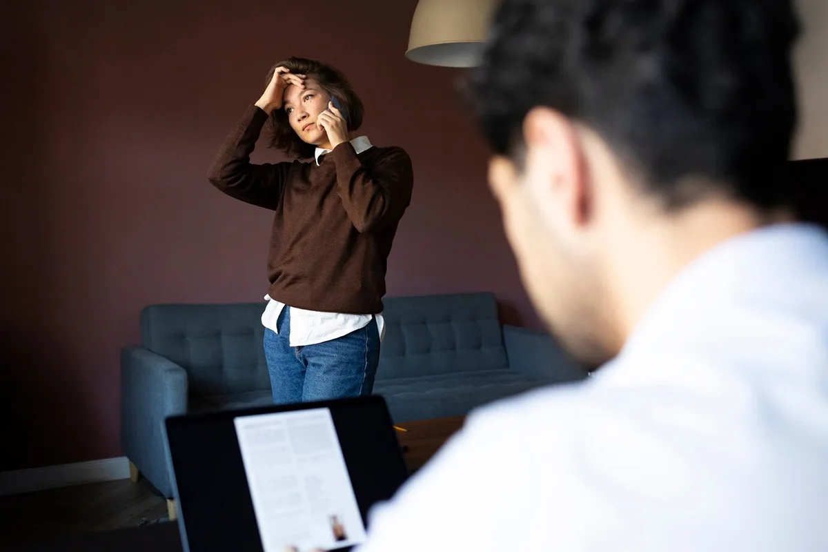 Woman on phone looking concerned, standing in living room near a man working on a laptop in foreground.