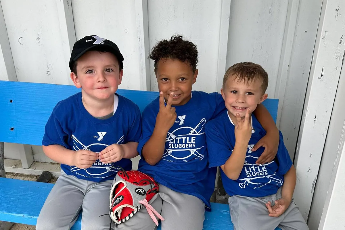 Three young boys in blue 'Little Sluggers' T-shirts sitting on a bench, smiling and flashing peace signs.