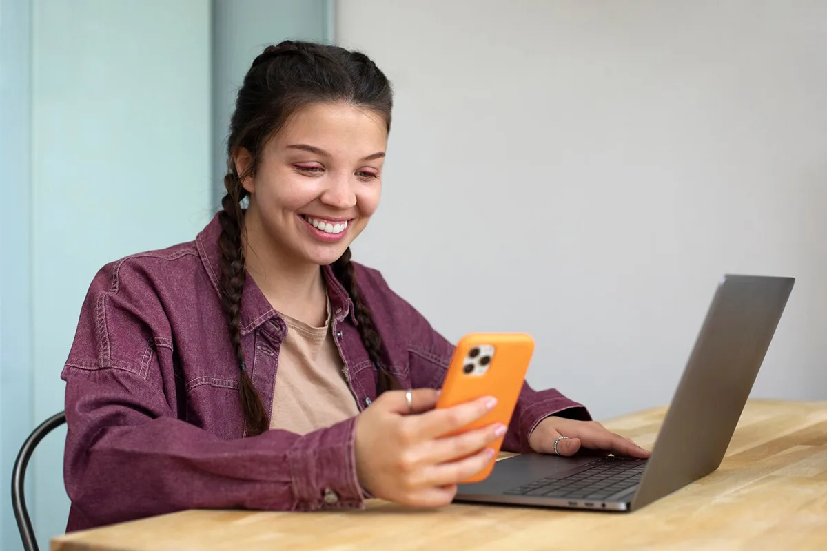 Smiling young woman using smartphone and laptop at a desk.