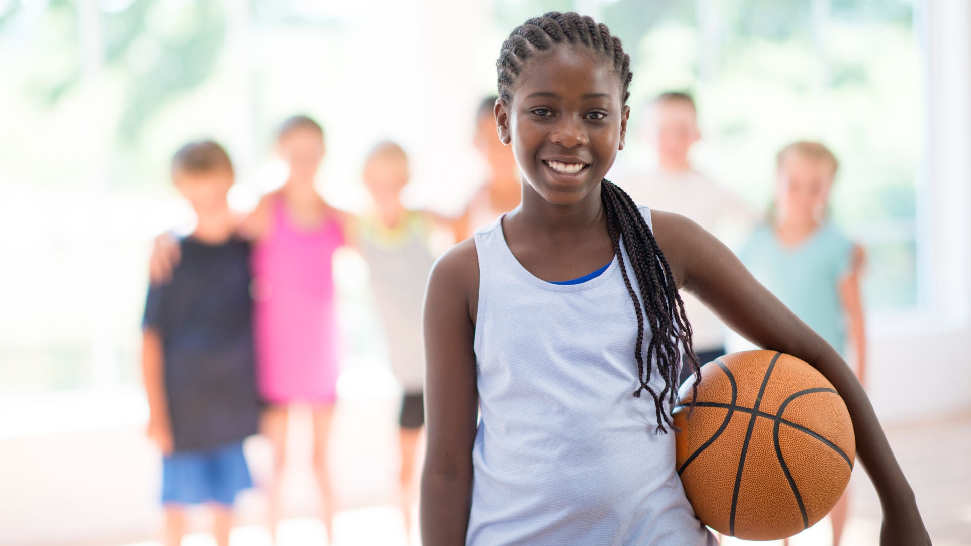 Smiling girl holding a basketball with a group of children standing blurred in the background in a brightly lit gym.