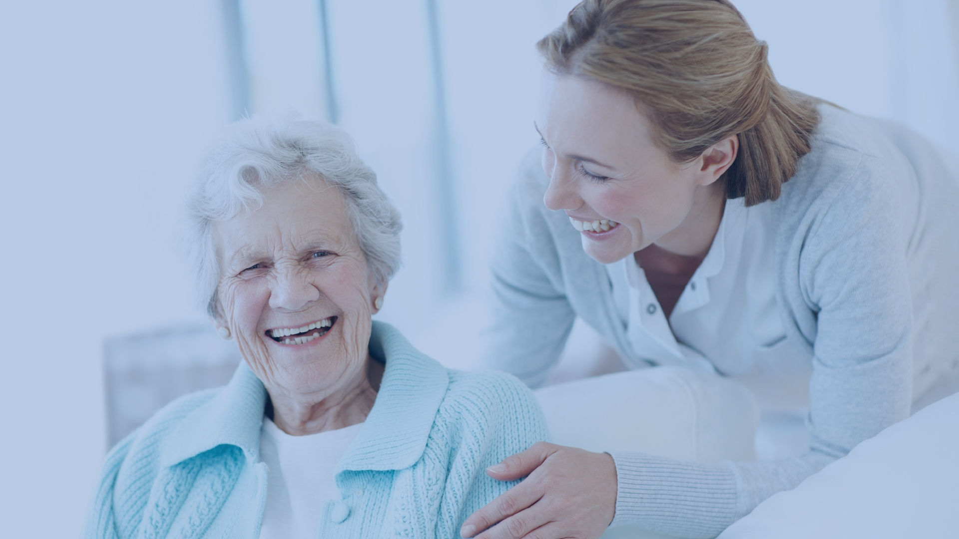 Smiling elderly woman sitting while a younger woman leans beside her, both sharing a joyful moment in a bright, cozy setting.