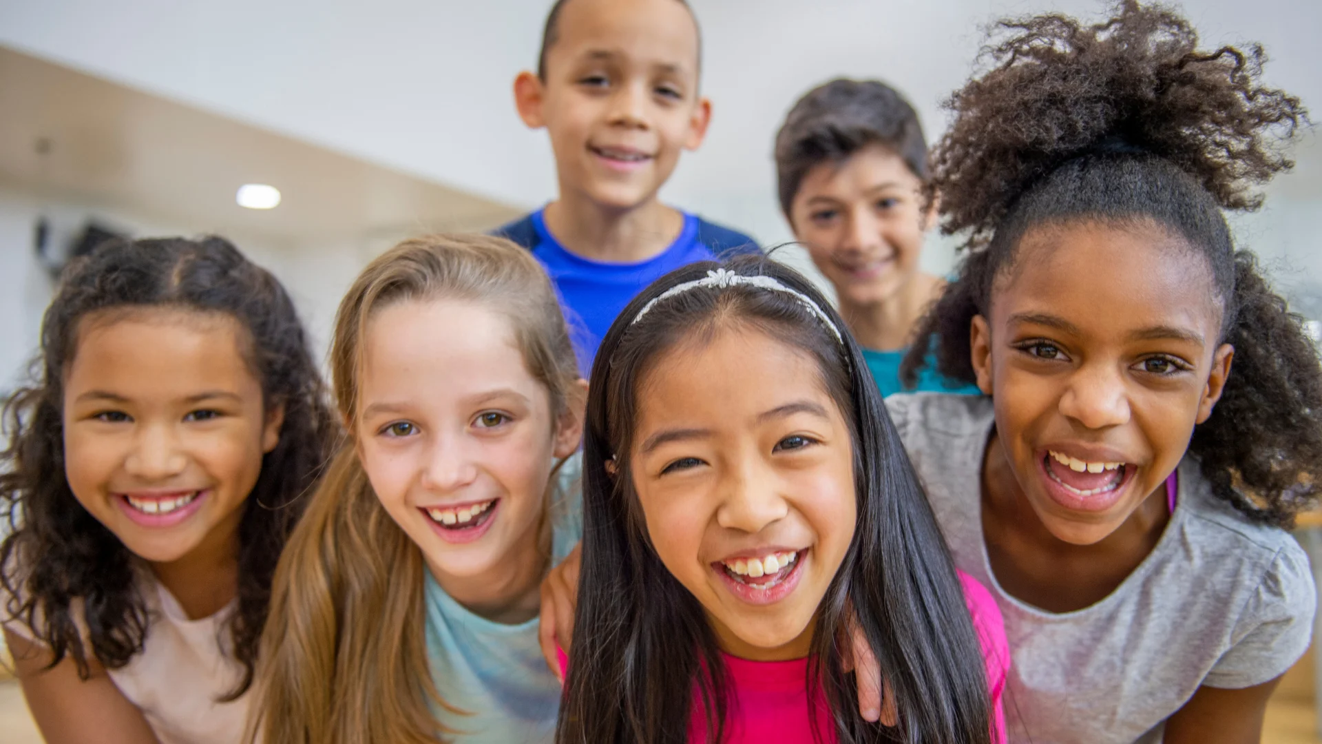 Group of smiling children leaning together in a brightly lit room, enjoying a fun moment.