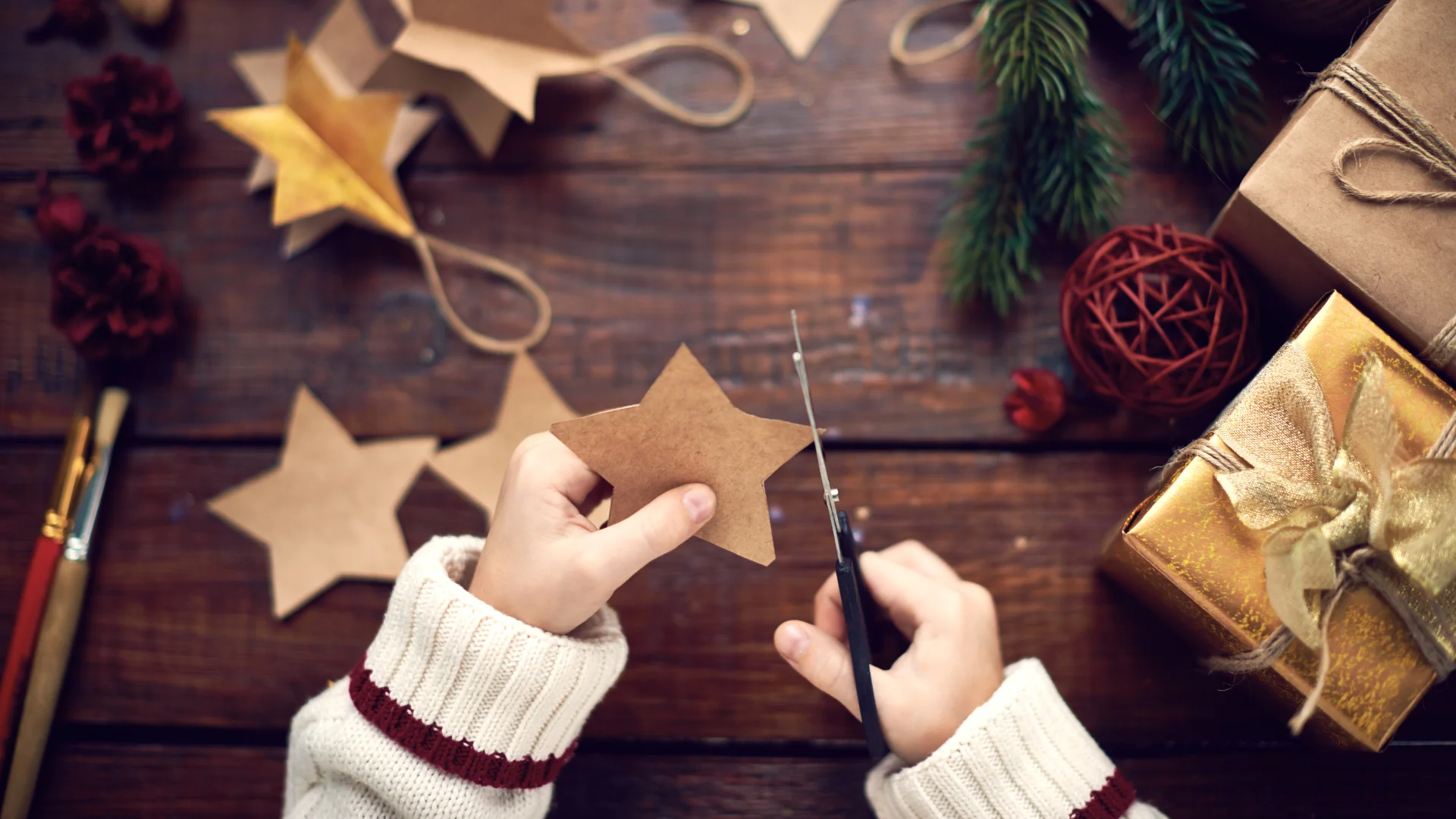 Person cutting a star-shaped ornament from brown paper on a wooden table decorated with craft supplies, pinecones, and wrapped gifts.