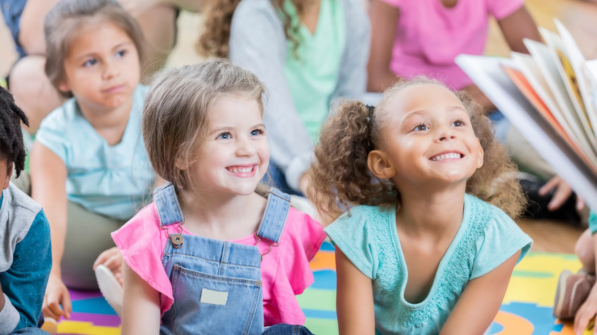 Group of young children sitting on a colorful mat, smiling and listening to a story being read aloud.