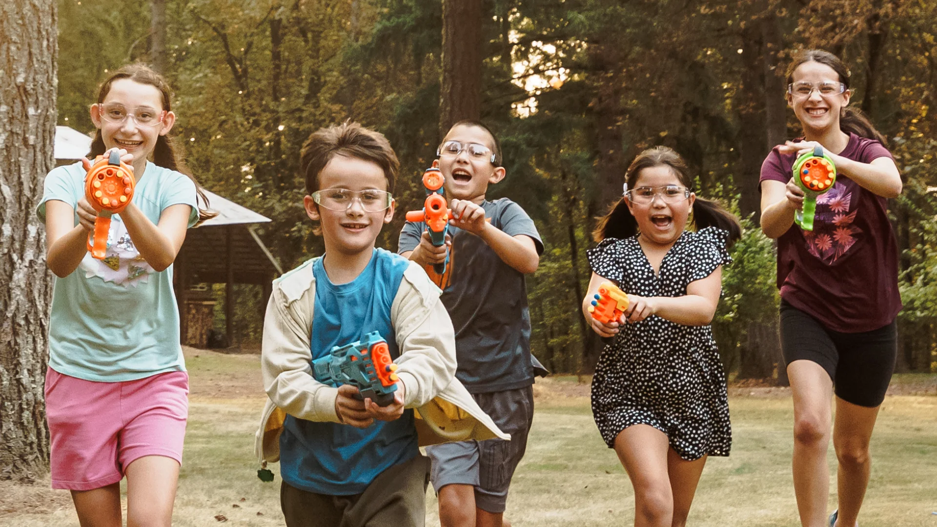 Group of children wearing safety goggles running outdoors and playing with toy blasters in a wooded area.