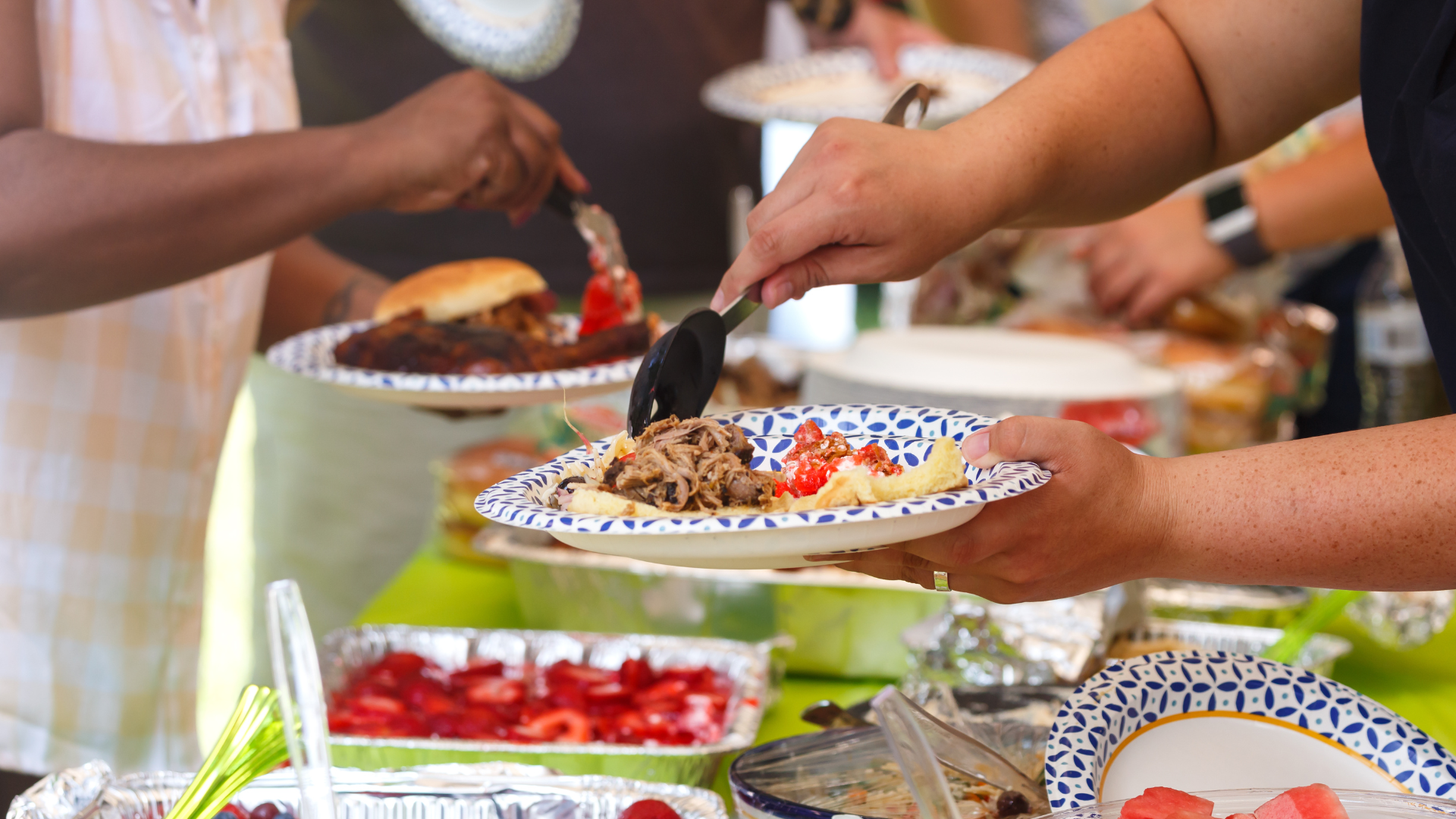 People serving food onto plates from a buffet table filled with dishes, including barbecue, salads, and desserts.