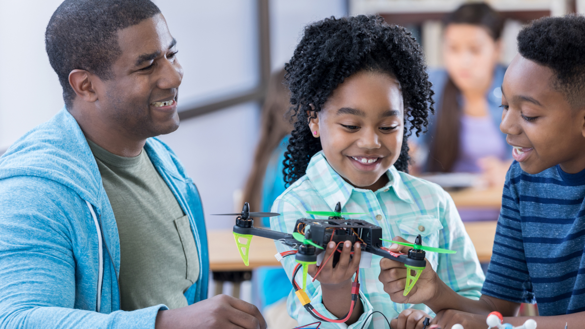 Smiling teacher guiding two students as they examine and hold a small drone in a classroom setting.