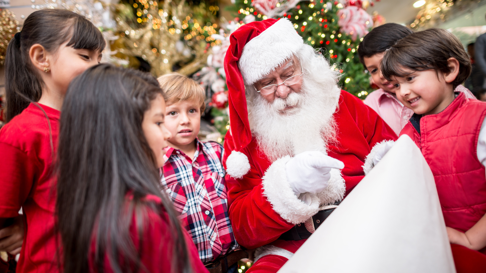 Santa Claus surrounded by children, reading a wish list in a festive setting with a decorated Christmas tree in the background.