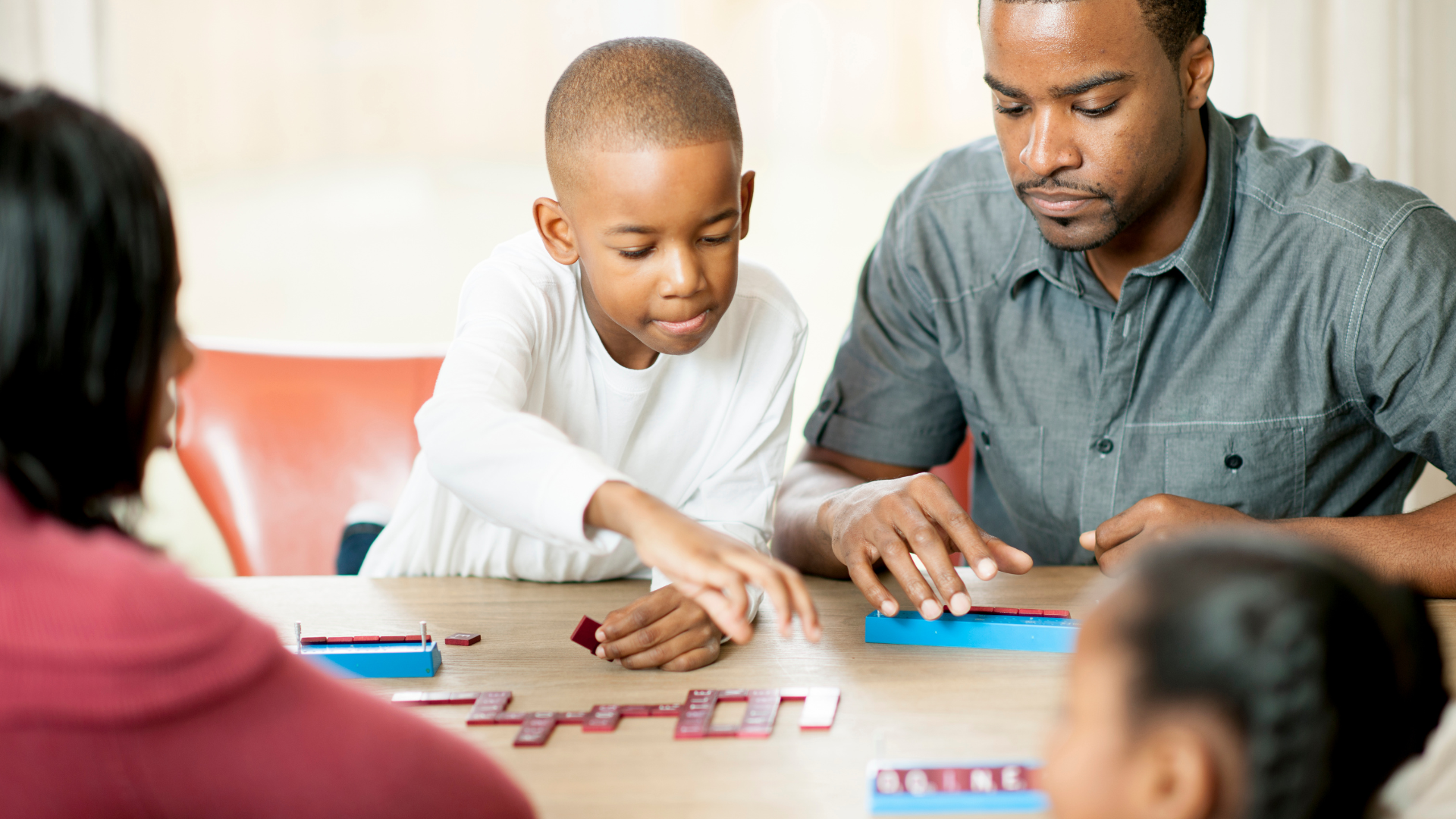 Family playing a board game together at a table, with a young boy making his move and an adult assisting.